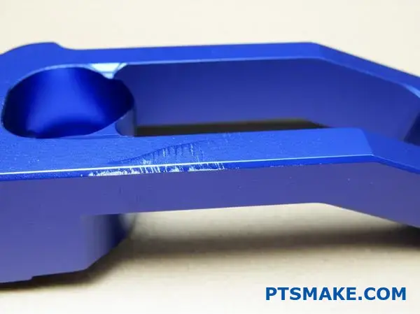 Detailný pohľad na modro eloxovaný hliníkový držiak s hlbokými škrabancami a mikrotrhlinami na povlaku